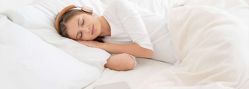 Eine junge Frau mit Kopfhörern schläft entspannt in einem Bett mit weißen Kissen und einer Decke.