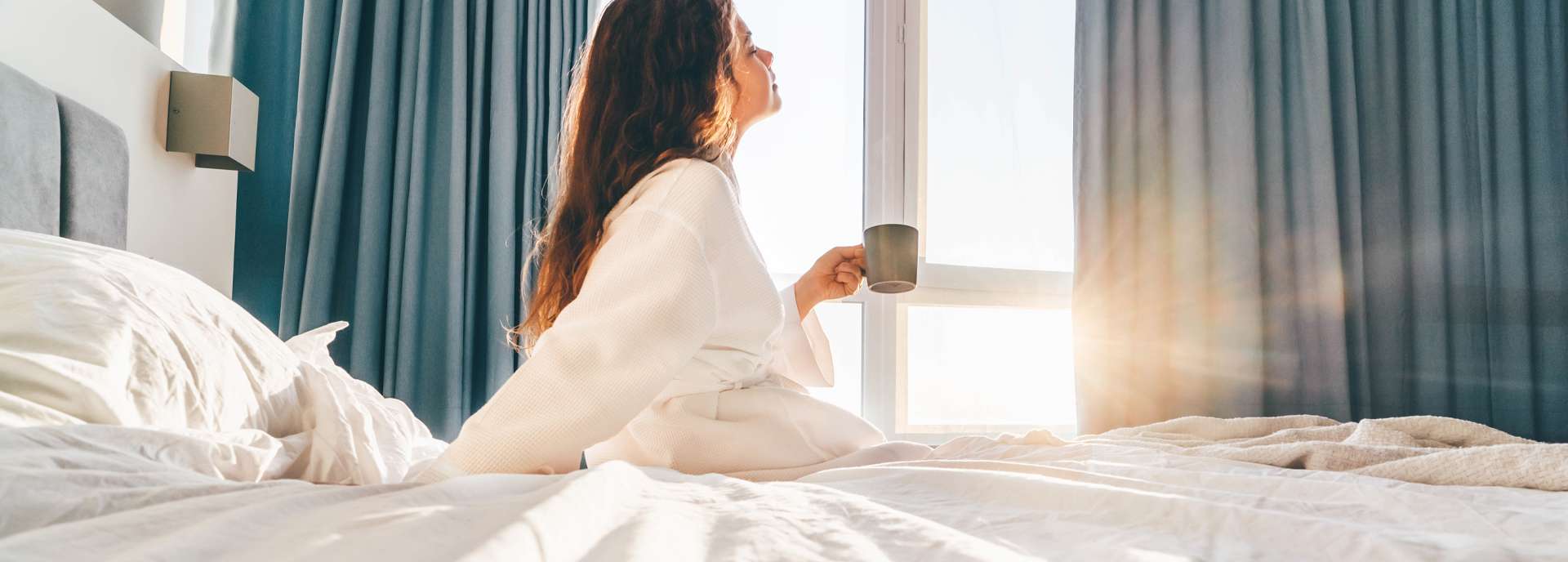 Frau in einem Morgenmantel sitzt auf dem Bett und genießt einen Kaffee beim Blick aus dem Fenster.