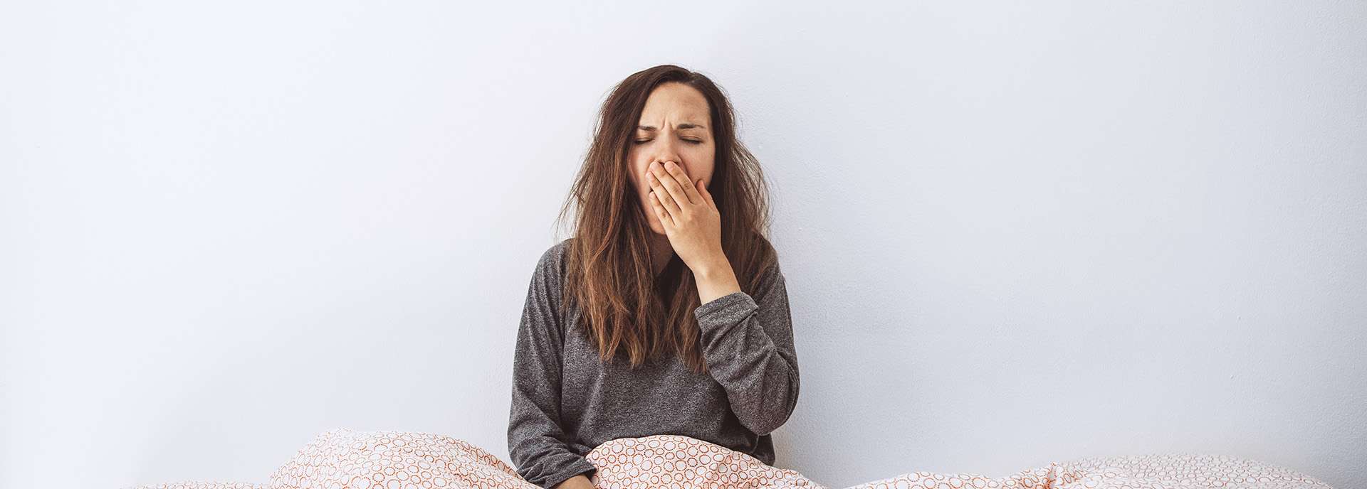 Frau mit zerzausten Haaren sitzt im Bett und gähnt, umgeben von einer Decke mit Muster.