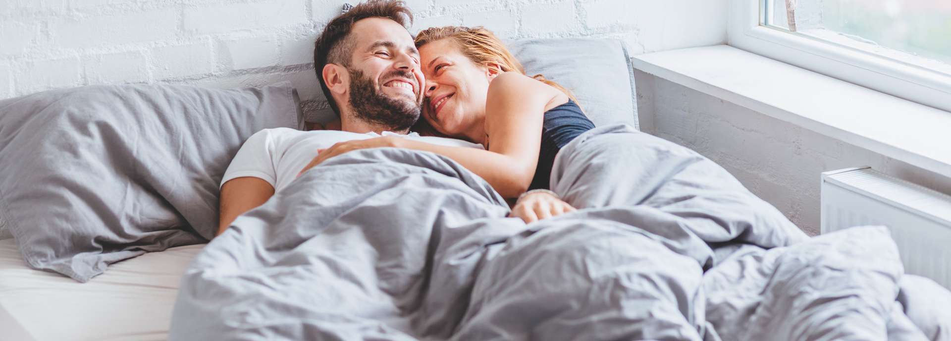 Paar liegt lächelnd im Bett unter einer Decke und genießt den gemeinsamen Moment. Tageslicht strömt durch das Fenster.