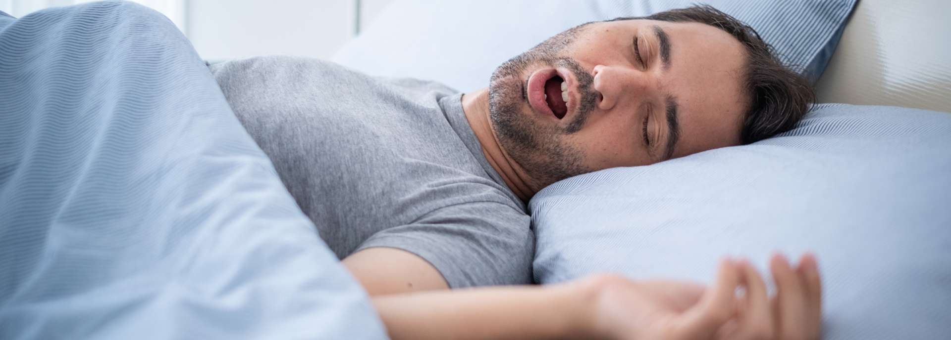 Mann schläft im Bett und schnarcht, mit offenem Mund und entspanntem Gesichtsausdruck.