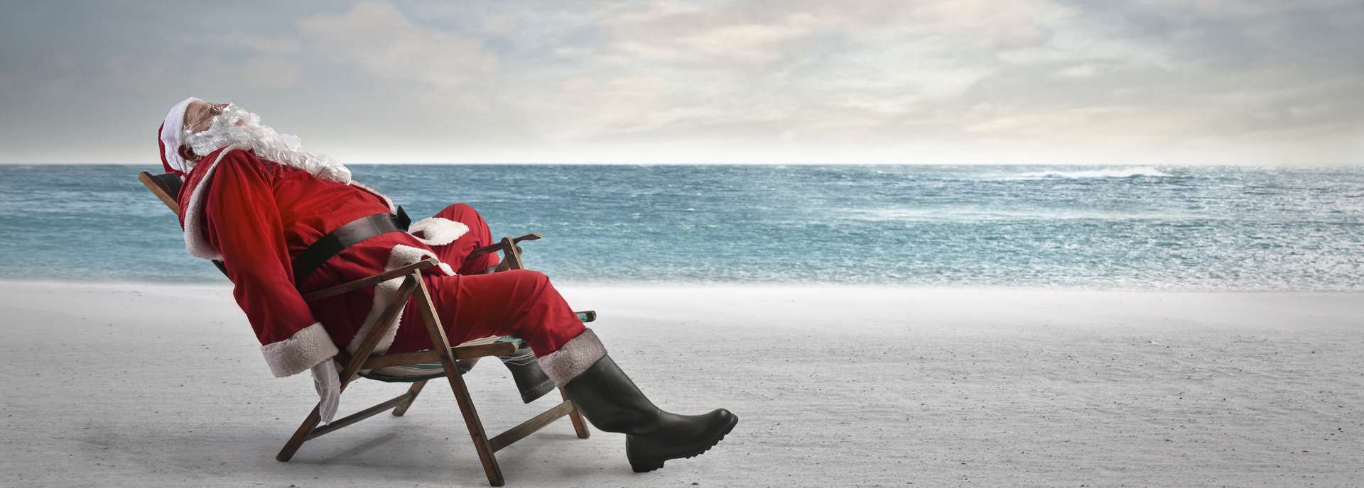 Ein Weihnachtsmann in rotem Anzug entspannt auf einem Liegestuhl am Strand mit Meerblick.
