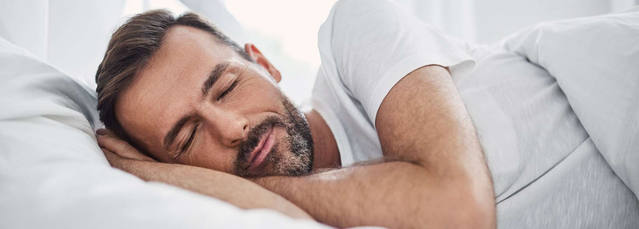 Mann schläft entspannt auf einem Bett mit Kopf auf einem Kissen, umgeben von weißen Bettwaren.