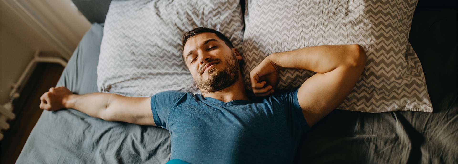 Mann liegt entspannt im Bett mit Kopf auf Kissen und Decke, umgeben von grauer Bettwäsche.
