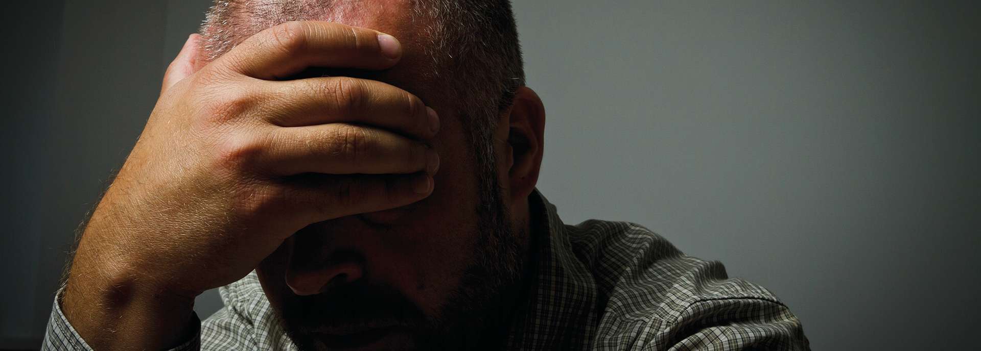 Mann mit gesenktem Kopf und Hand auf der Stirn, in einer dunklen Umgebung, wirkt nachdenklich oder gestresst.