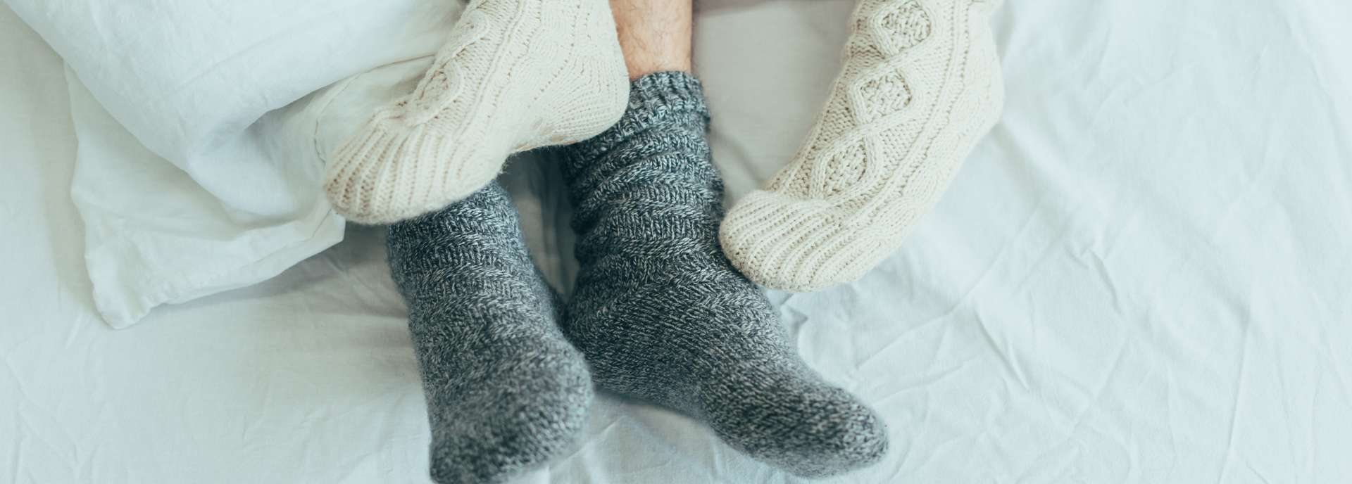 Füße in warmen Socken liegen unter einer Decke auf einem Bett mit weißer Bettwäsche.