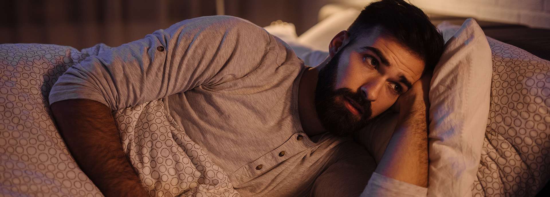 Mann liegt im Bett und schaut nachdenklich zur Decke, umgeben von Kissen und Decken.