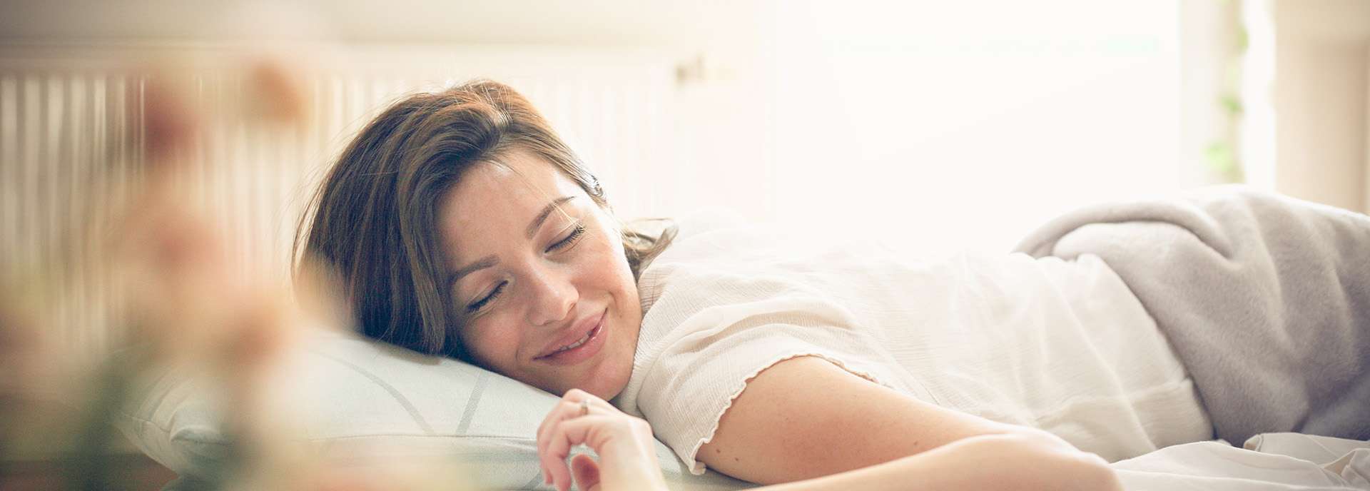 Eine Frau liegt entspannt im Bett und lächelt, umgeben von weicher Bettwäsche und Tageslicht.
