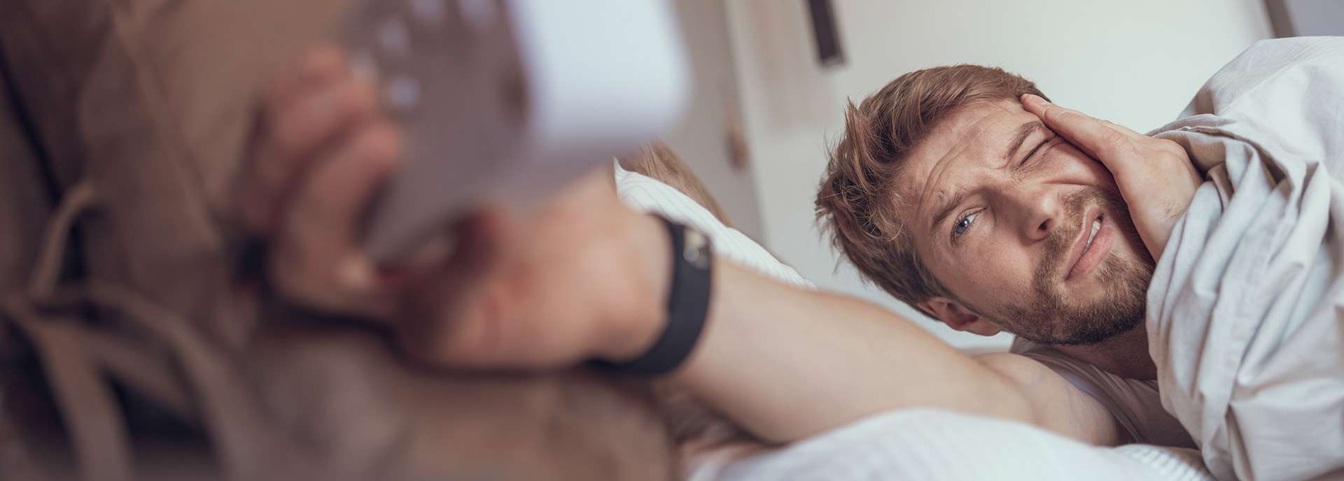 Mann im Bett mit Fernbedienung, sieht müde und verwirrt aus, während er auf die Uhr schaut.
