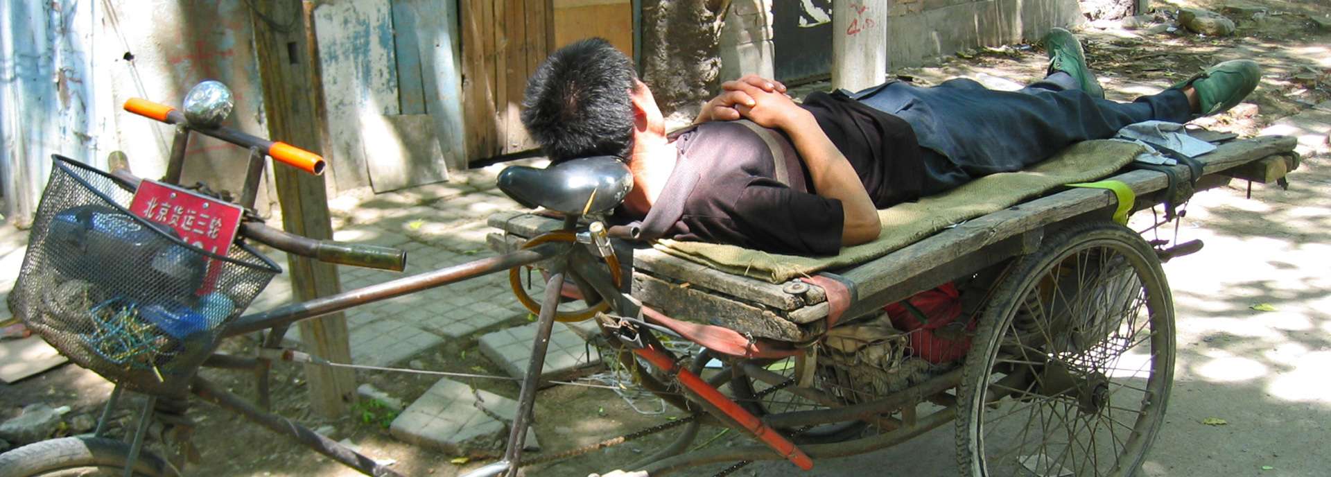 Mann liegt entspannt auf einem Handwagen in einer ruhigen Straße, umgeben von Bäumen und Gebäuden.