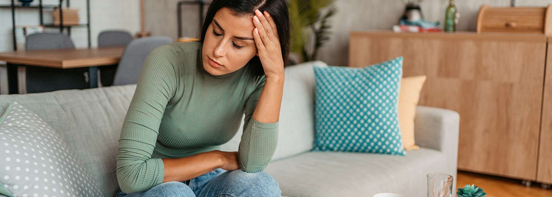 Frau sitzt nachdenklich auf einer Couch, hält sich die Stirn und wirkt müde in einem modernen Wohnraum.