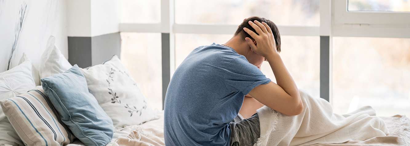 Mann sitzt auf einem Bett, hält den Kopf in den Händen und blickt nach unten, umgeben von Kissen und einer Decke.