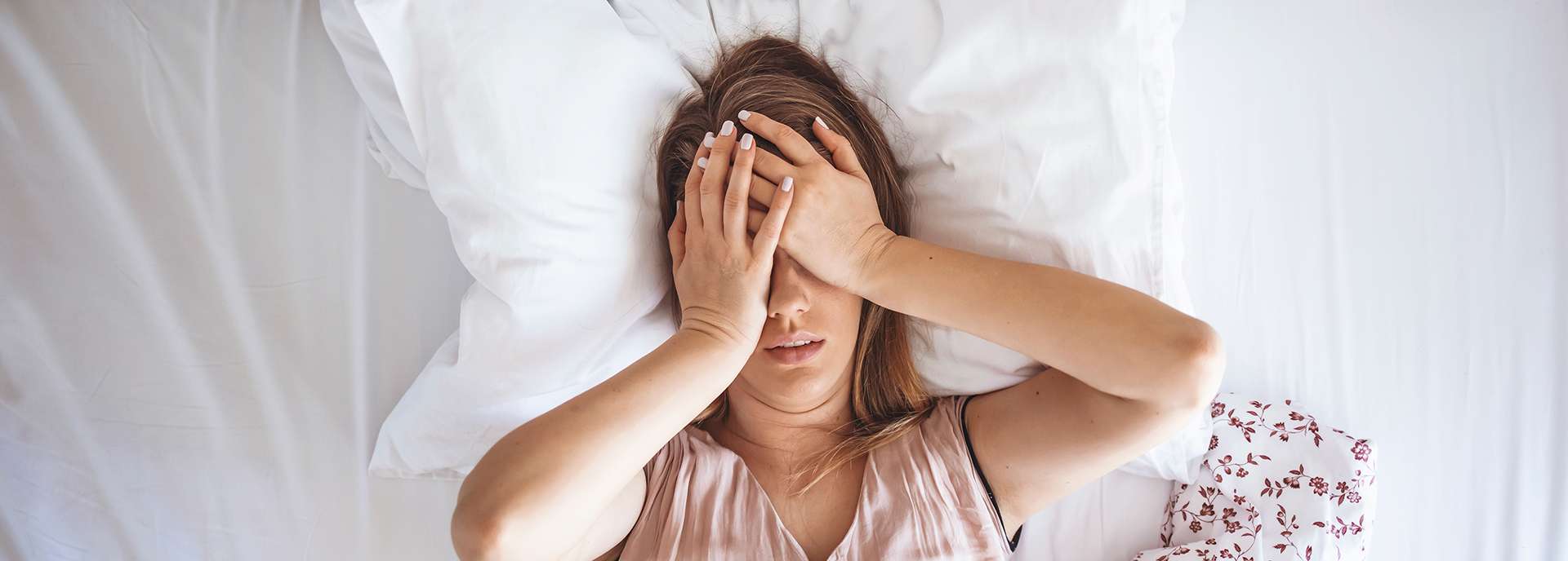 Frau liegt im Bett mit den Händen im Gesicht, wirkt gestresst oder unruhig. Kissen und Bettdecke sichtbar.