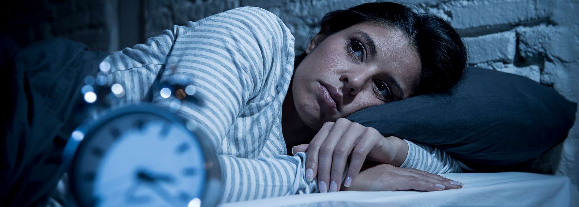 Frau liegt wach im Bett, schaut nachdenklich zur Seite, Wecker im Vordergrund zeigt die späte Stunde.