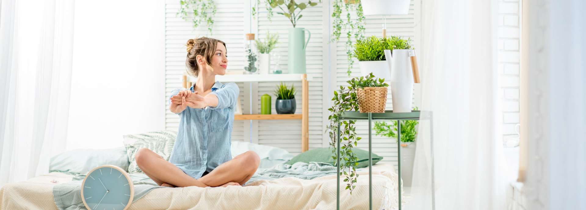 Frau sitzt im Schneidersitz auf einem Bett, umgeben von Pflanzen und dekorativen Elementen in einem hellen Raum.