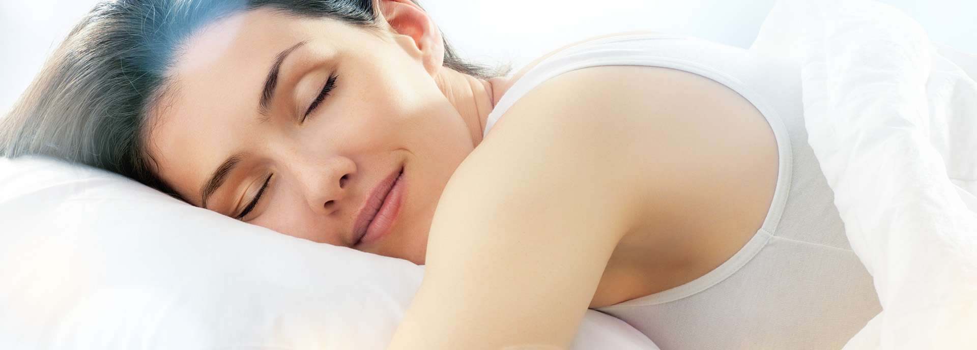 Frau schläft entspannt auf einem Kissen, umgeben von weichen Bettlaken und sanftem Licht.