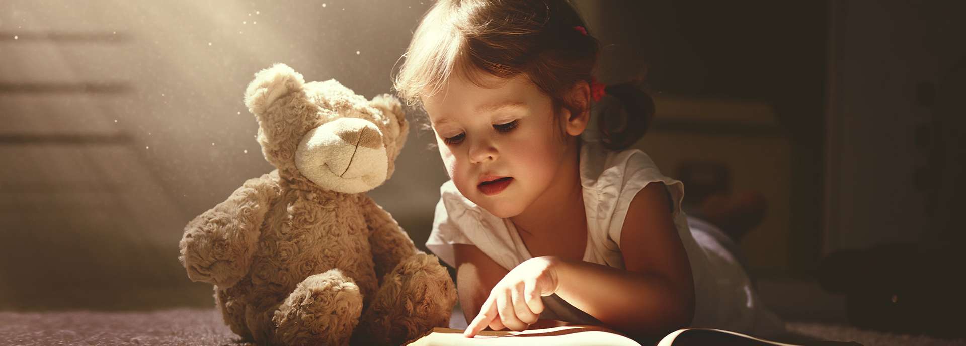 Ein Kind liegt auf dem Boden und liest ein Buch, neben ihm steht ein plüschiger Bär. Sanftes Licht fällt ins Bild.
