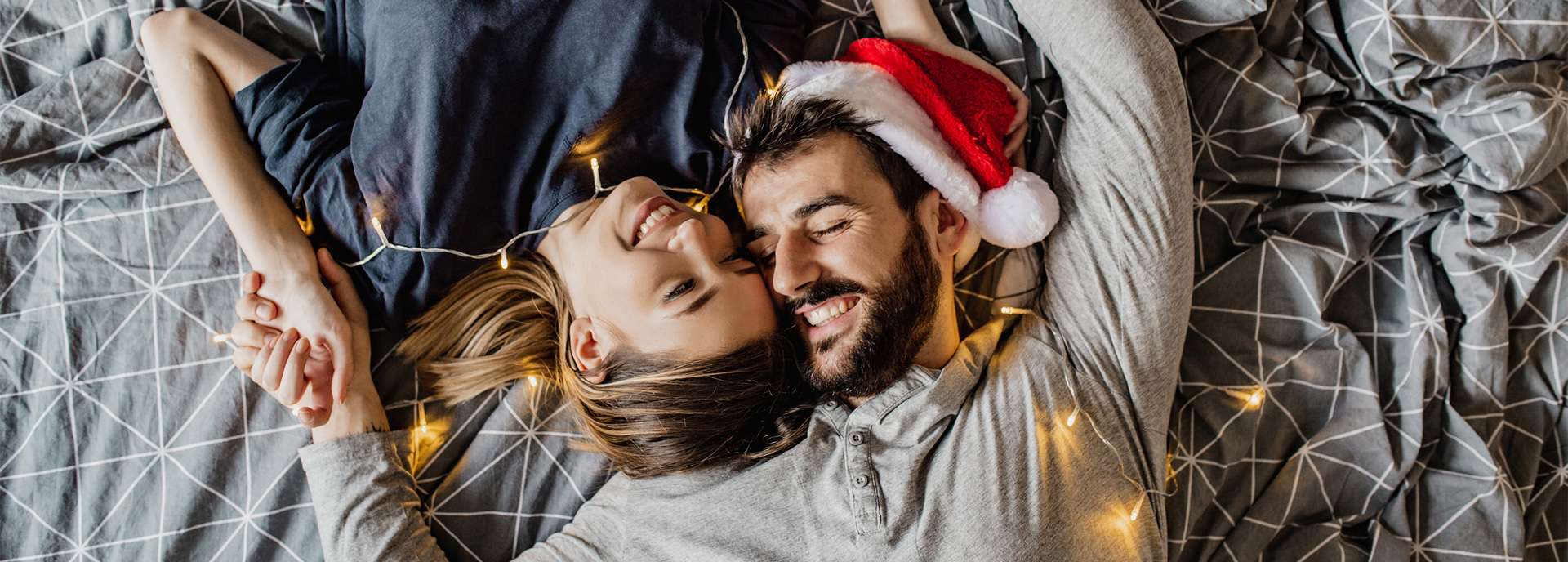 Paar liegt auf einem Bett, umgeben von Lichtern, mit Weihnachtsmütze, lächelnd und entspannt.