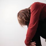 Donna seduta su uno sgabello, si china in avanti abbracciandosi le ginocchia, appare pensierosa e triste.