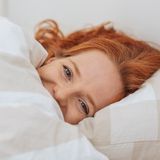 Donna sorridente con capelli rossi, sdraiata a letto sotto una coperta a righe.