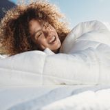 Frau mit lockigem Haar, die glücklich in eine Bettdecke eingekuschelt auf einem Bett liegt.