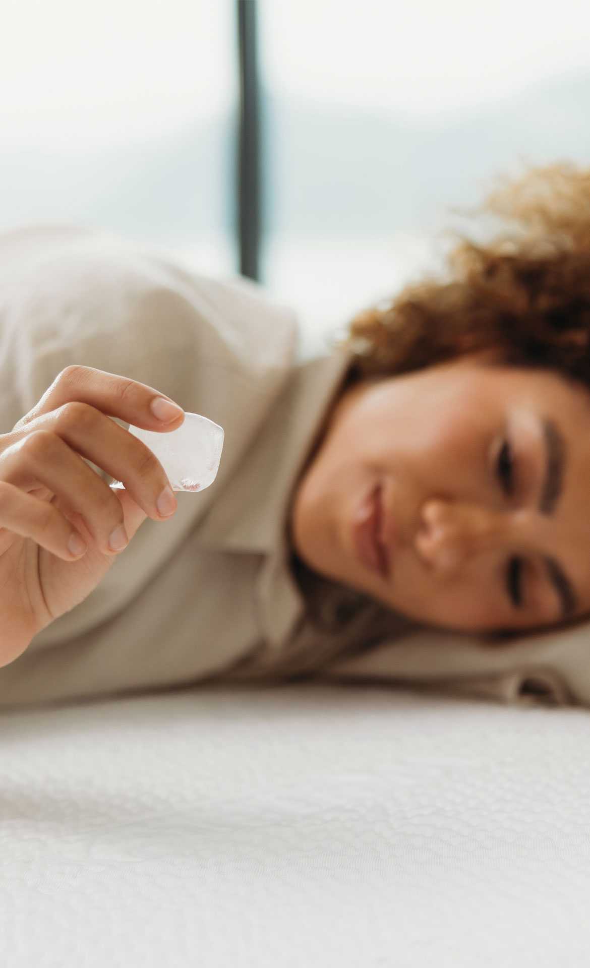 A person lies on a mattress holding an ice cube in their hand.