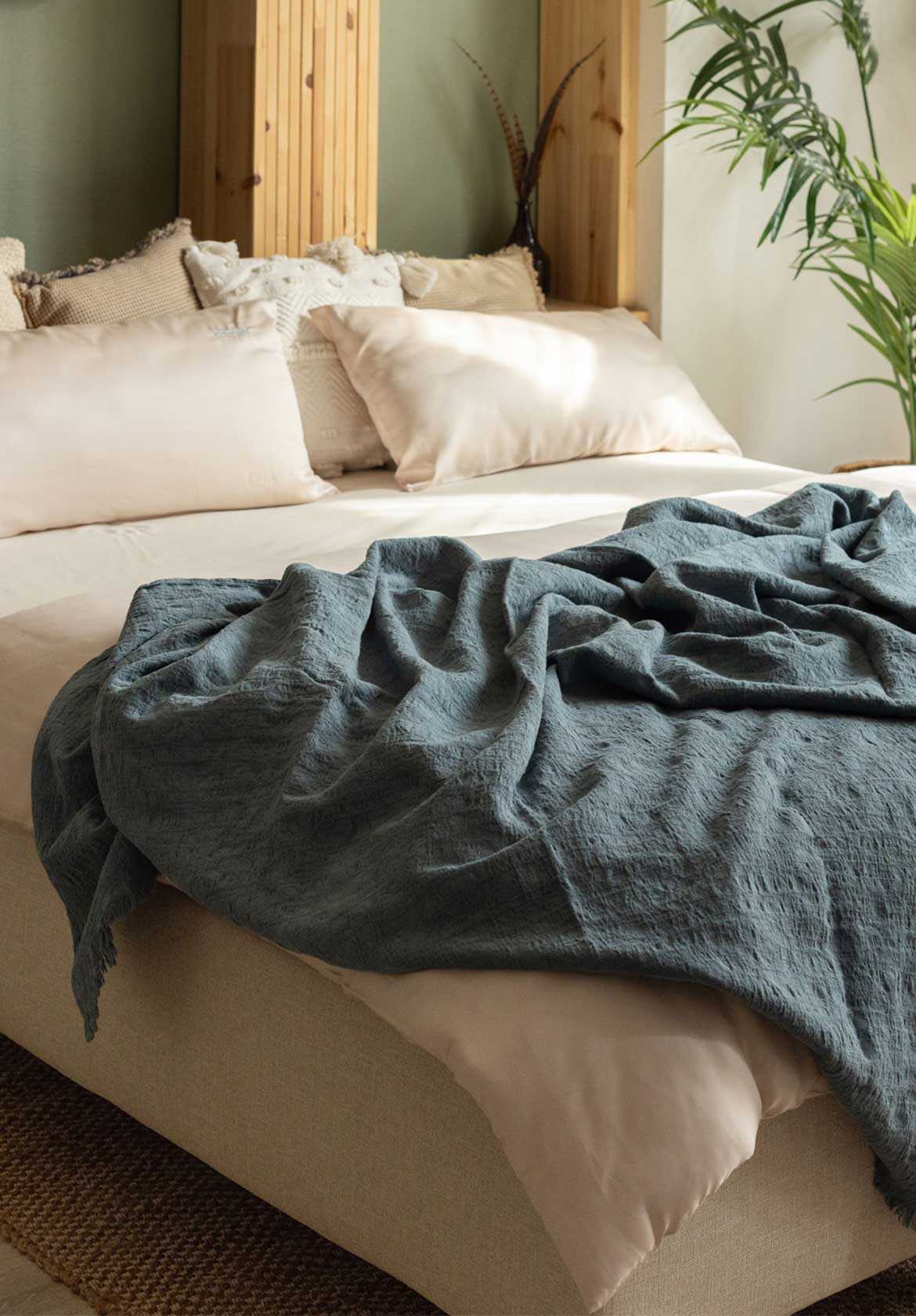 Bett mit hellen Kissen und einer blauen Decke, umgeben von Pflanzen und Holzdetails im Hintergrund.