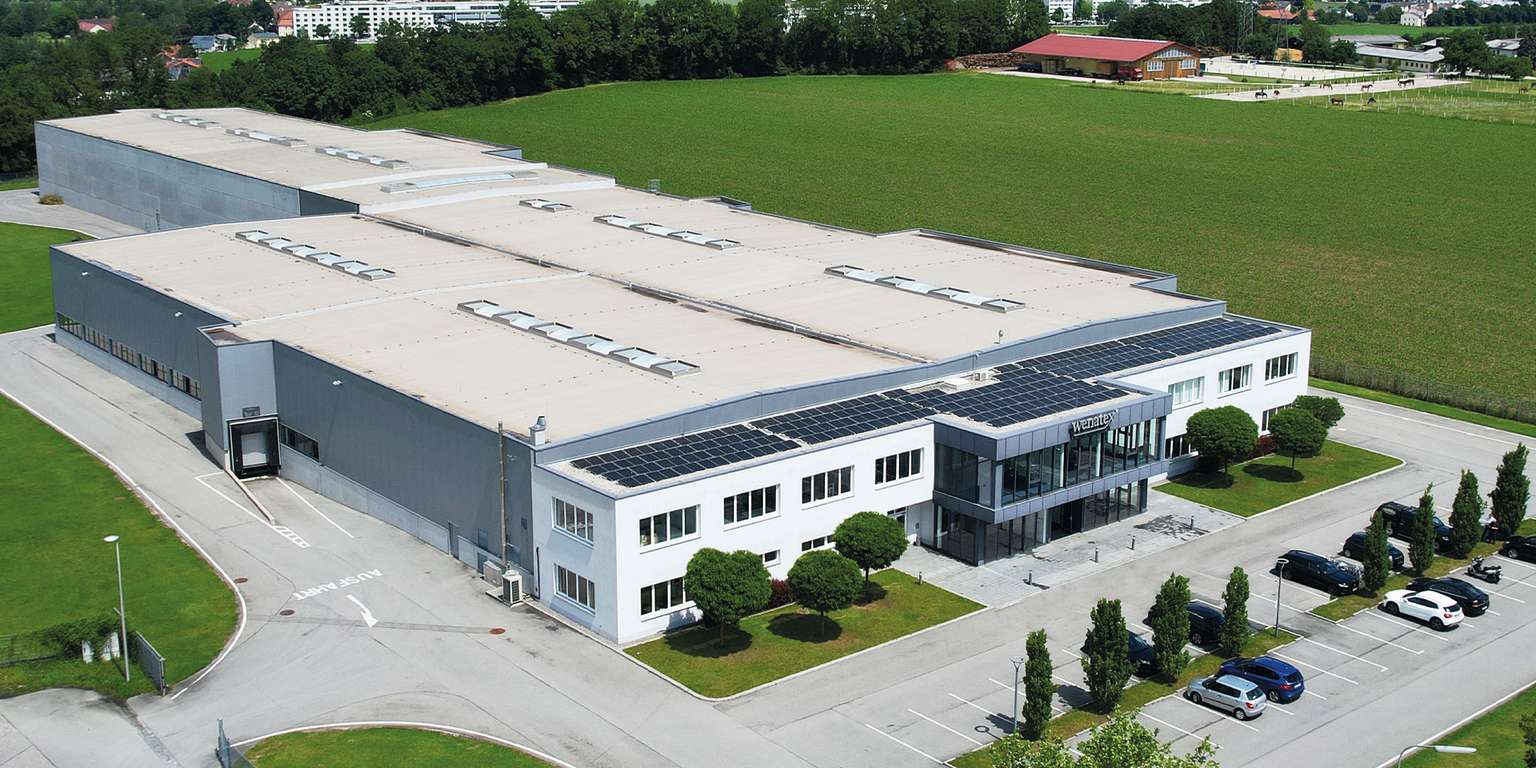 Modern production hall with solar panels on the roof, surrounded by green spaces and parking lots.