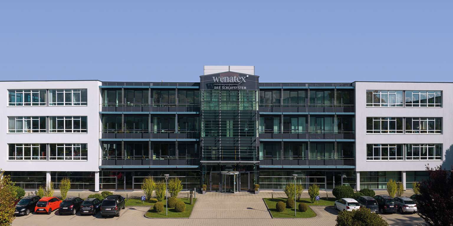 Moderne Bürogebäude mit Glasfassade und Parkplatz, umgeben von Bäumen und Grünflächen.
