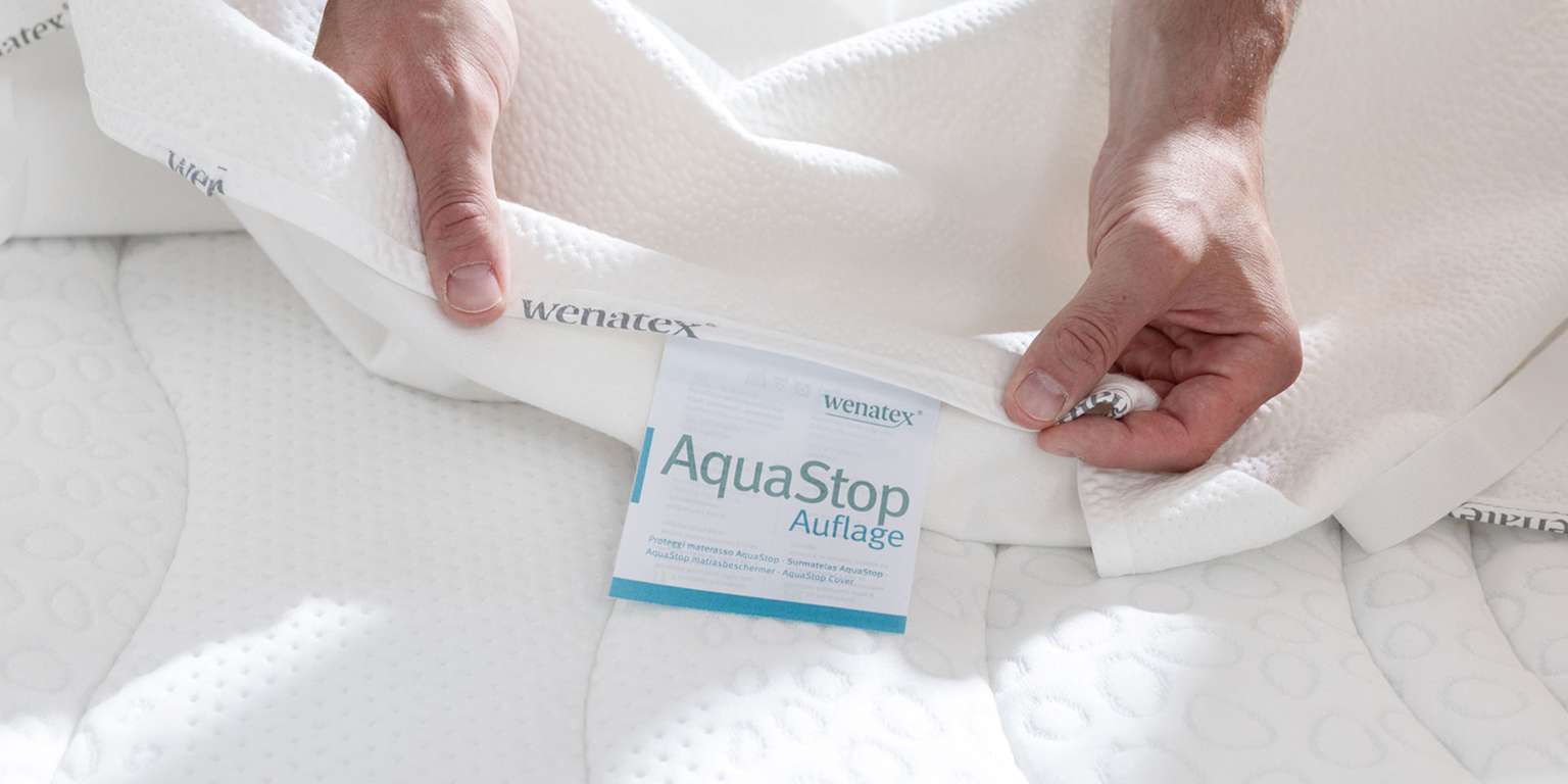 Hände halten eine Wenatex AquaStop Auflage, die auf einer Matratze platziert ist.