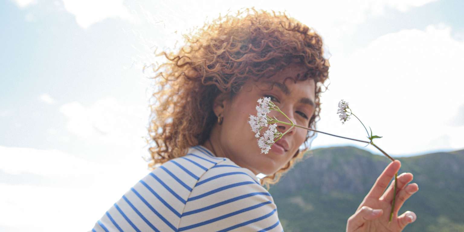 Eine Frau mit lockigem Haar hält eine Blume vor ihr Gesicht, umgeben von einer natürlichen Landschaft.