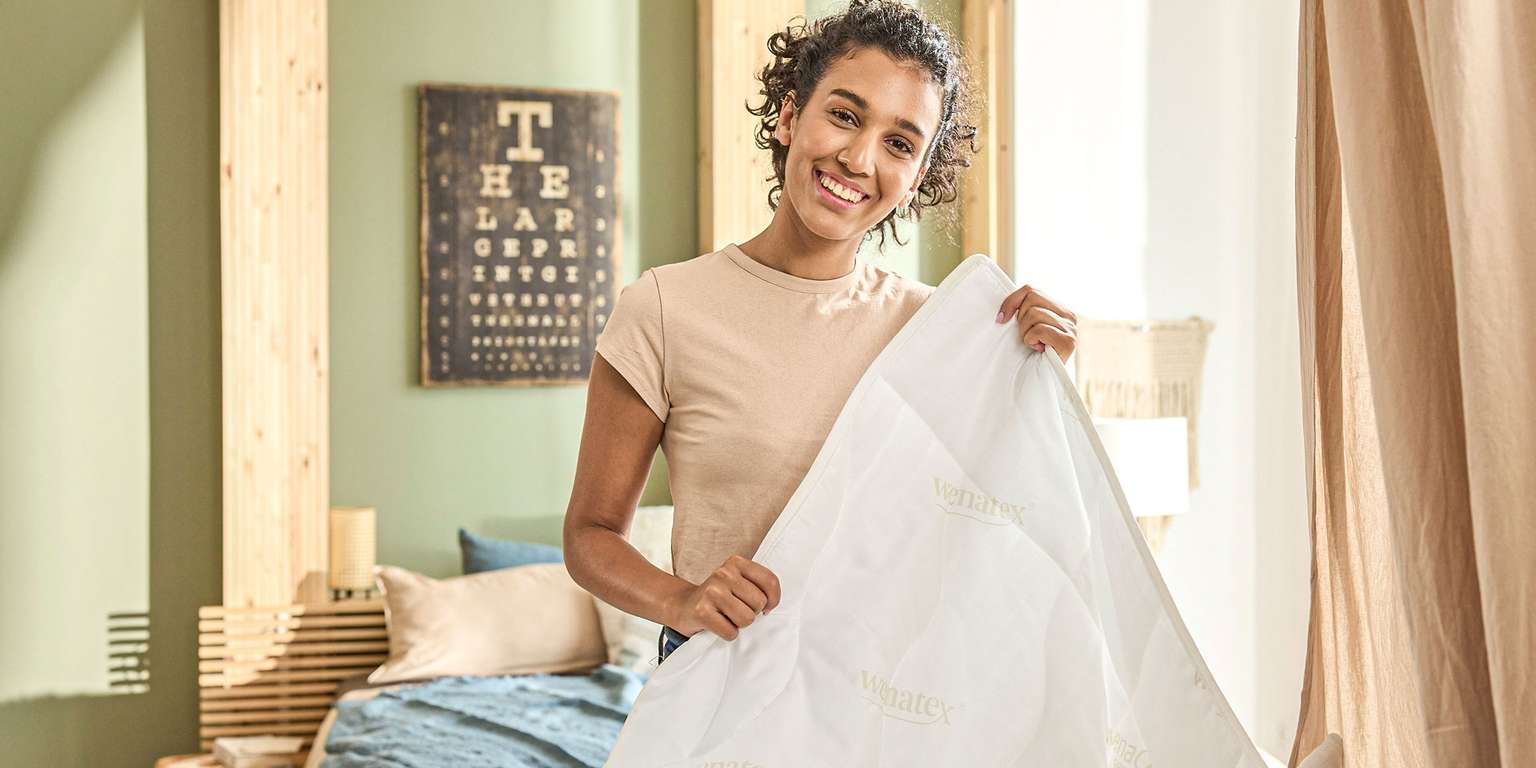 Frau hält eine Bettdecke von Wenatex in einem modernen Schlafzimmer mit Bett und dekorativen Elementen.