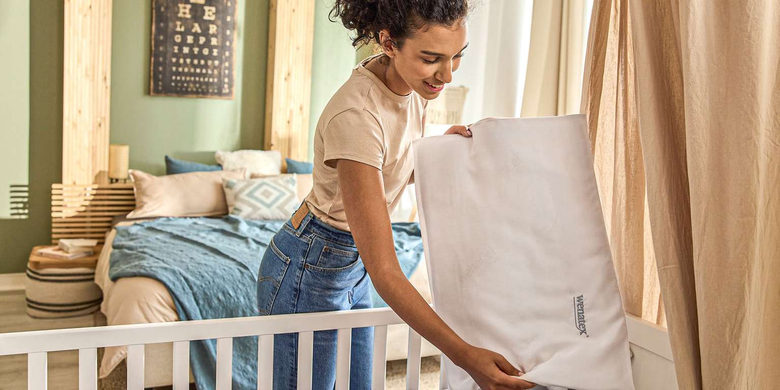 Frau legt ein Kissen in ein Babybett, umgeben von einem modernen Schlafzimmer mit sanften Farben.