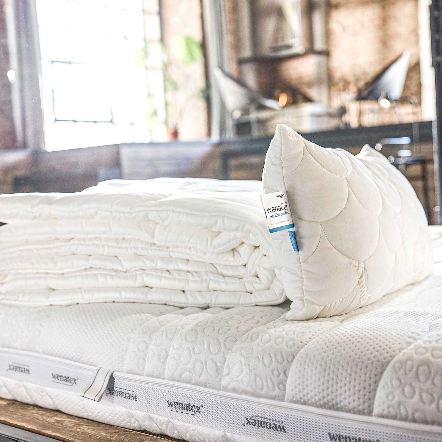 Matratze mit Matratzenauflage und Kissen von Wenatex auf einem Bett in einem modernen Raum.