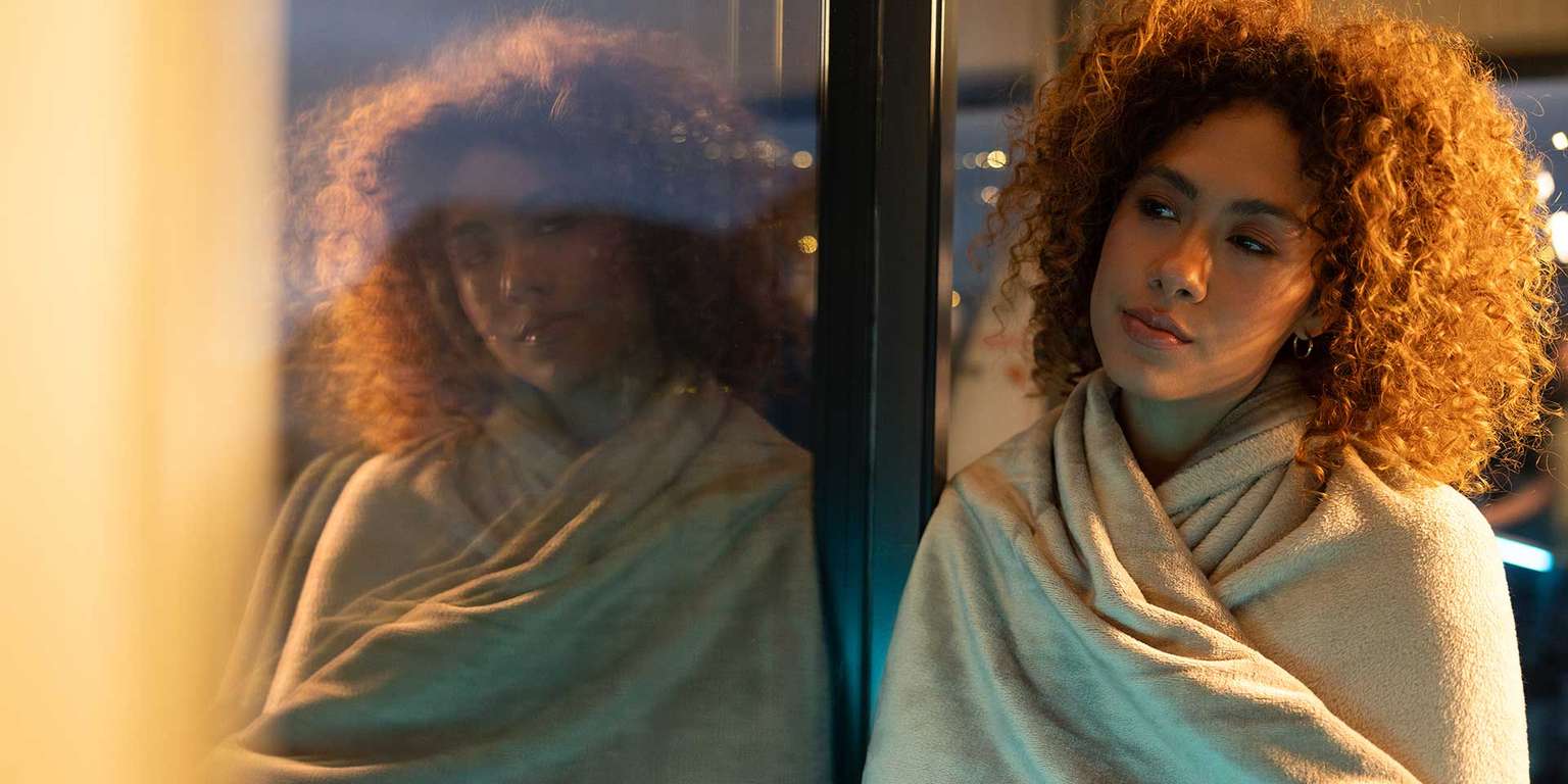 Frau mit lockigem Haar, die in eine kuschelige Decke gehüllt vor einem Fenster sitzt.