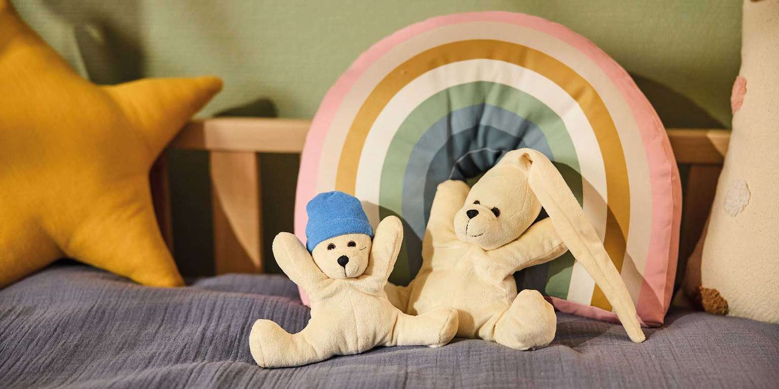 Zwei Stoffbären auf einem Bett mit buntem Kissen und einer farbigen Regenbogen-Dekoration.