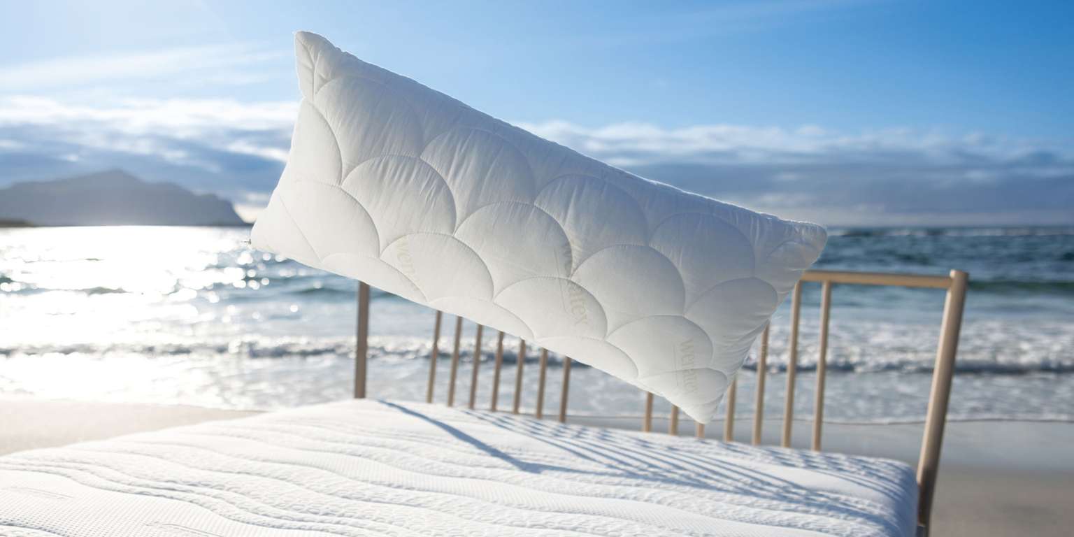 Wenatex Kissen schwebt über einem Bett am Strand mit Meerblick und sonnigem Himmel.