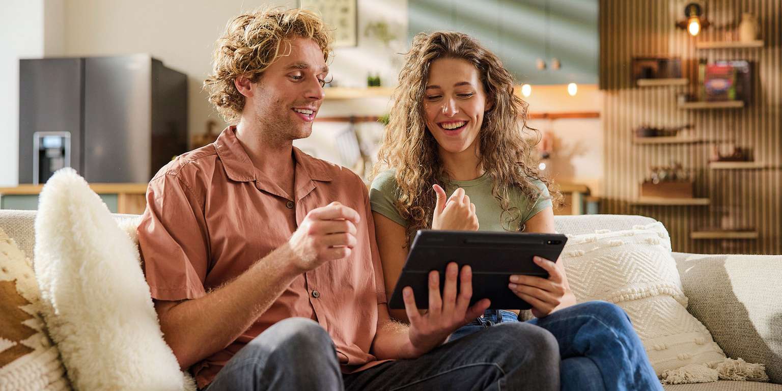 Couple sitting on a couch together looking at a tablet, both smiling and giving a thumbs up.