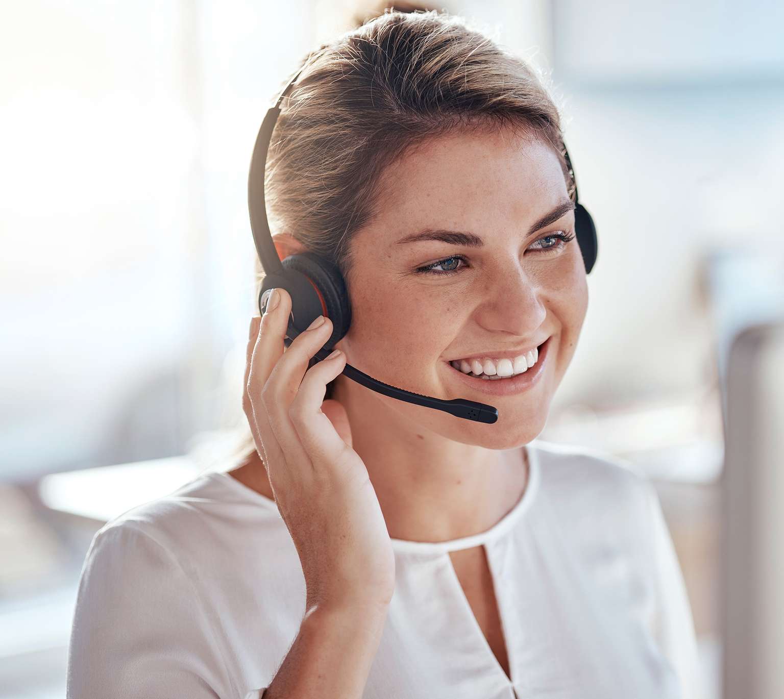 Frau mit Headset lächelt und spricht, in einem modernen Büro mit natürlichem Licht.