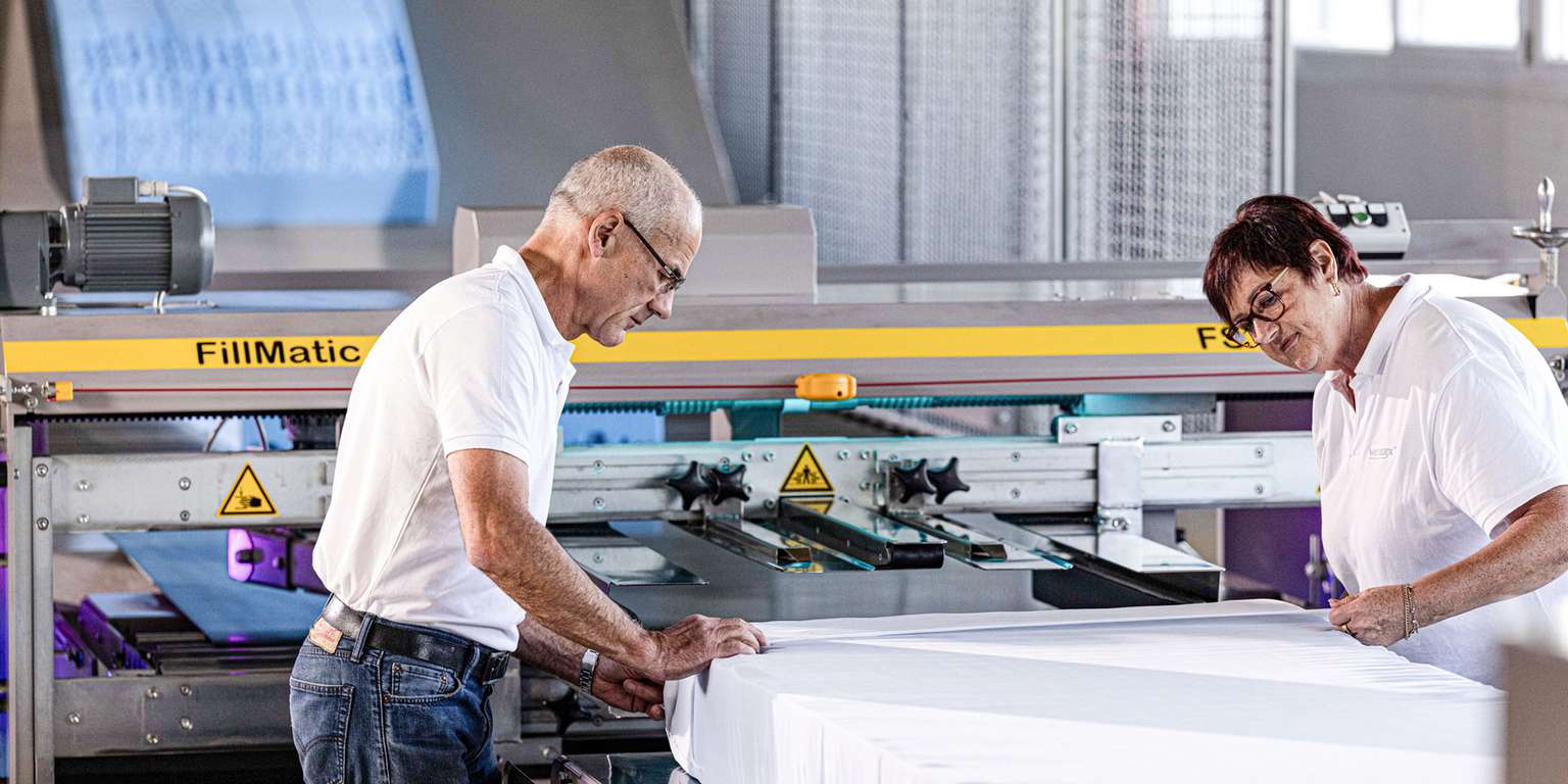 Zwei Personen arbeiten an einer Maschine zur Herstellung von Matratzenbezügen in einer Produktionsumgebung.
