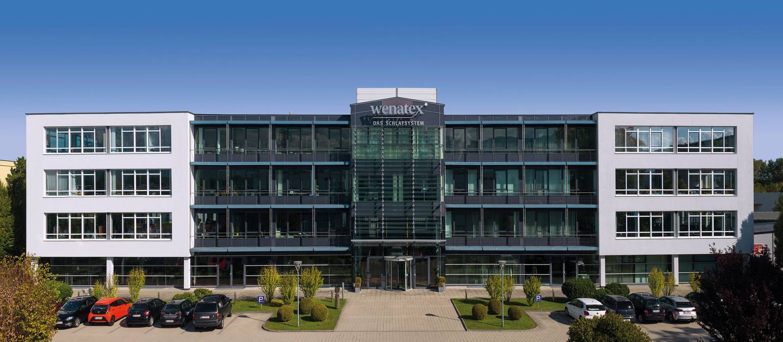 Moderne Bürogebäude mit Glasfassade, umgeben von Bäumen und parkenden Autos. Klarer blauer Himmel im Hintergrund.