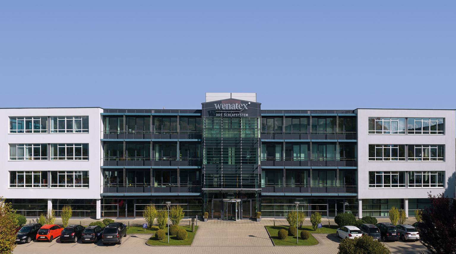 Modern office buildings with glass facades and parking, surrounded by trees and shrubs.