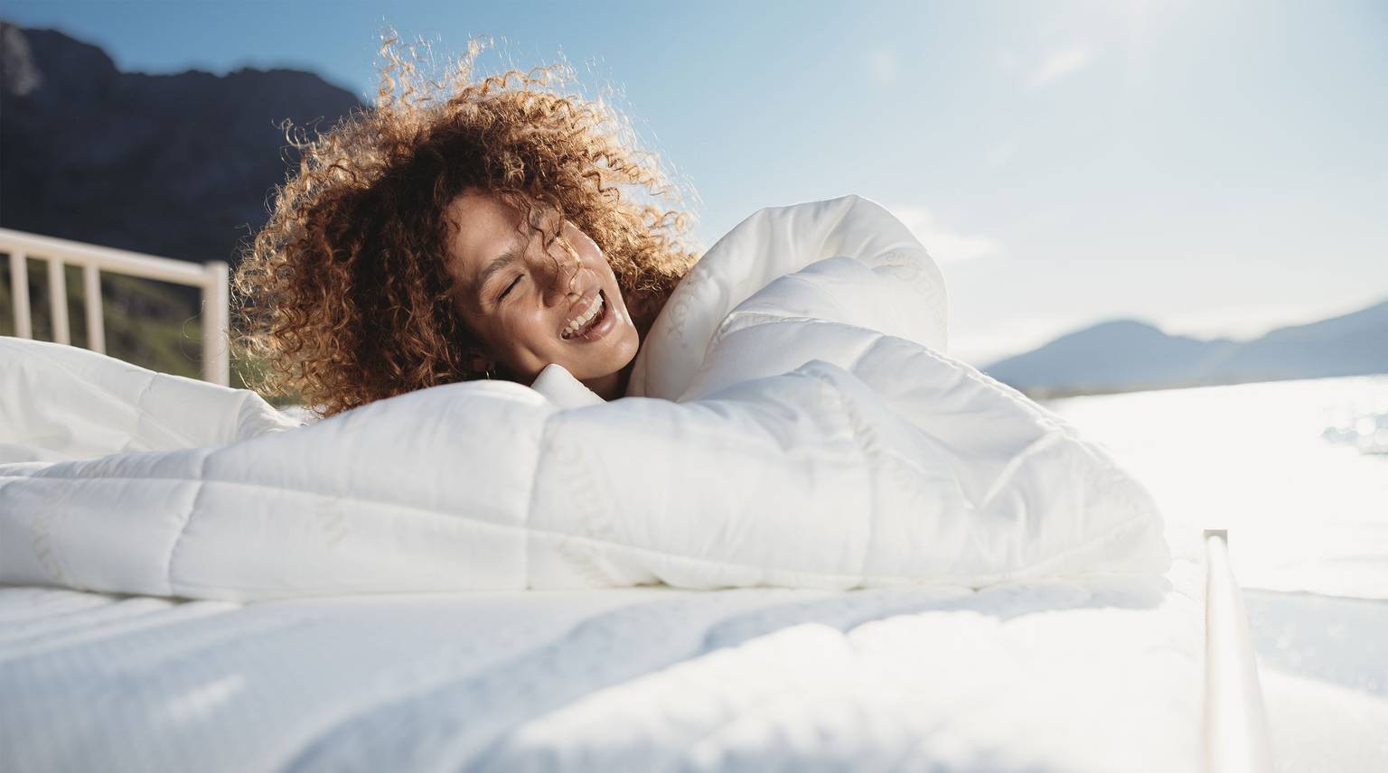 Frau mit lockigem Haar, die glücklich in eine Bettdecke eingekuschelt auf einem Bett liegt.