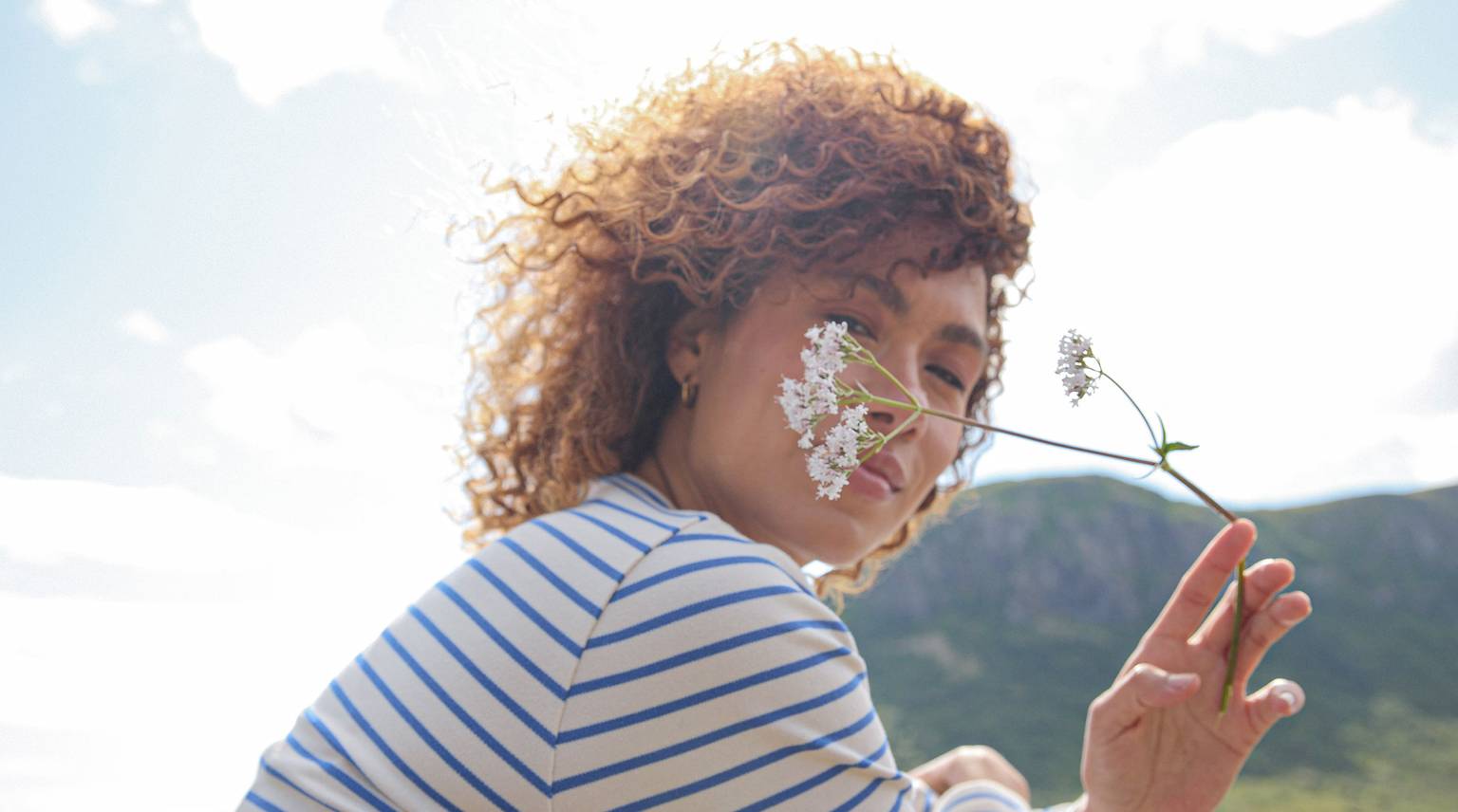 A woman with curly hair holds a flower in front of her face, surrounded by natural landscape.