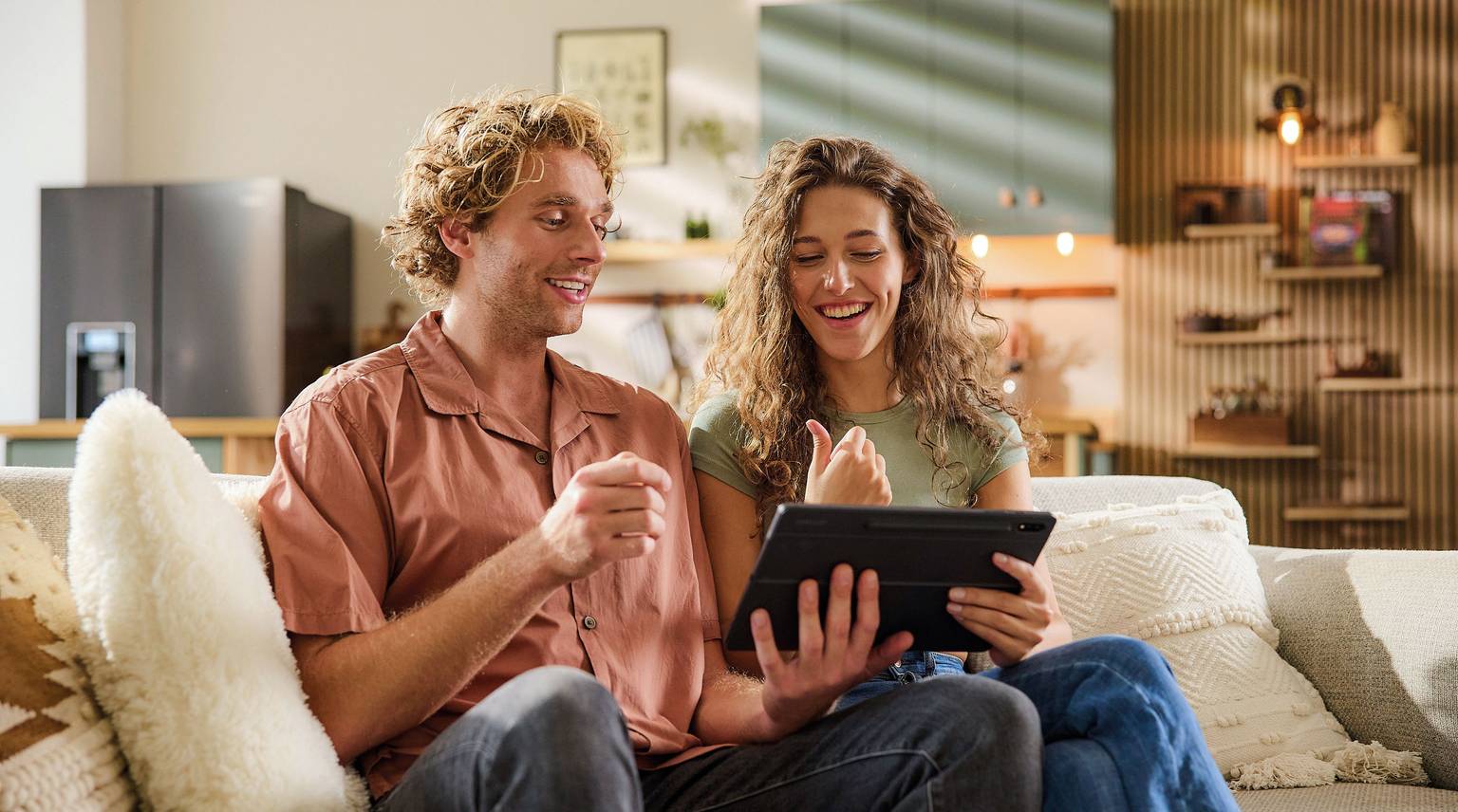 Couple sitting on a couch, smiling and giving a thumbs up while looking at a tablet together.