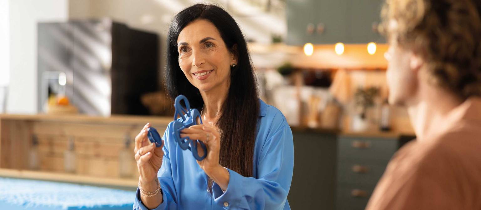 Eine Frau in blauer Bluse zeigt einem Mann in braunem Shirt ein Paar Schlafbrillen in einem hellen Raum.