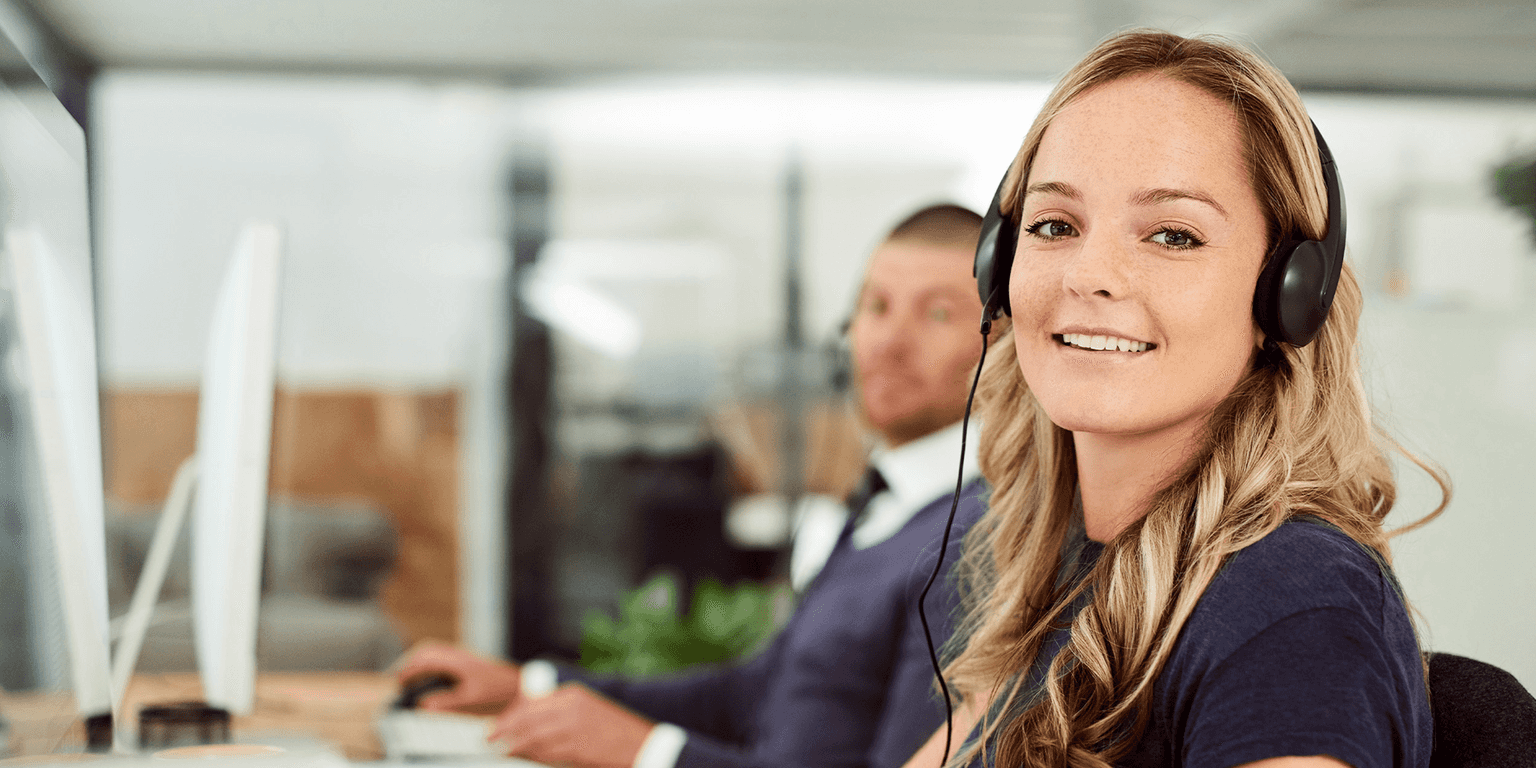 Lächelnde Frau mit Headset in einem modernen Büro, im Hintergrund ein Mann am Computer.
