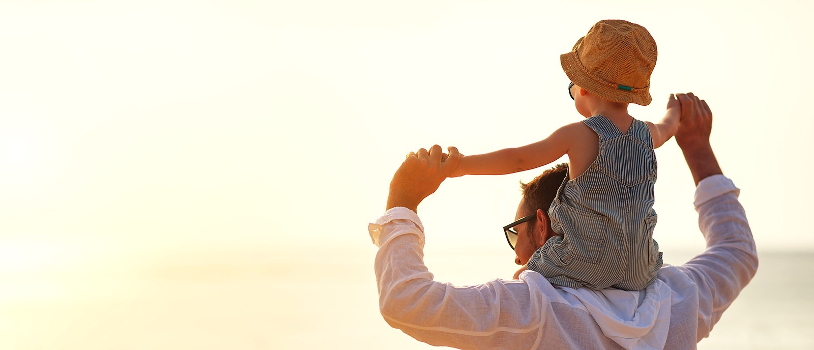Padre che porta il suo bambino sulle spalle in spiaggia, circondati da luce solare e atmosfera rilassata.