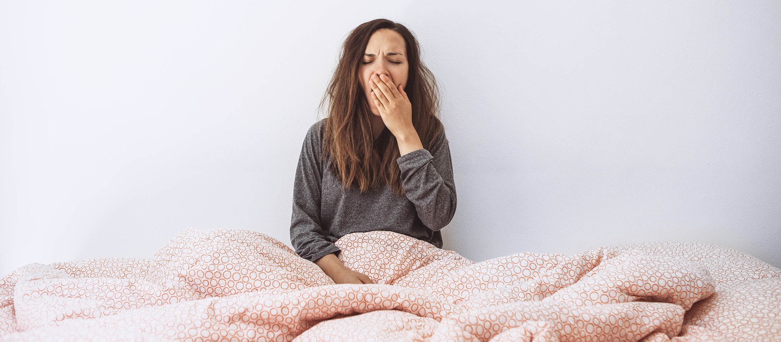 Frau sitzt im Bett, gähnt und wirkt müde, umgeben von einer Decke mit einem geometrischen Muster.