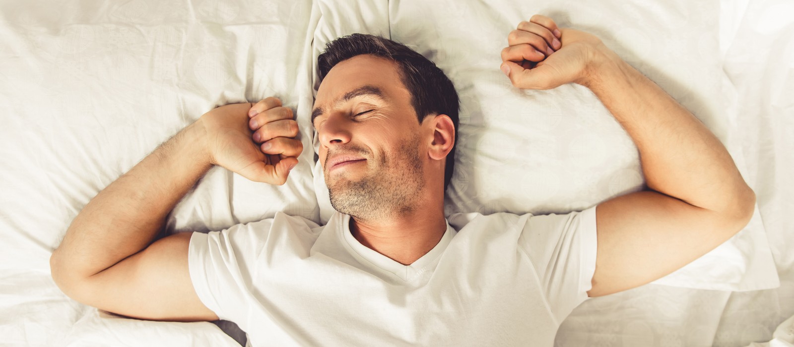 Uomo rilassato su un letto, sorride e si stende in una camera da letto luminosa.